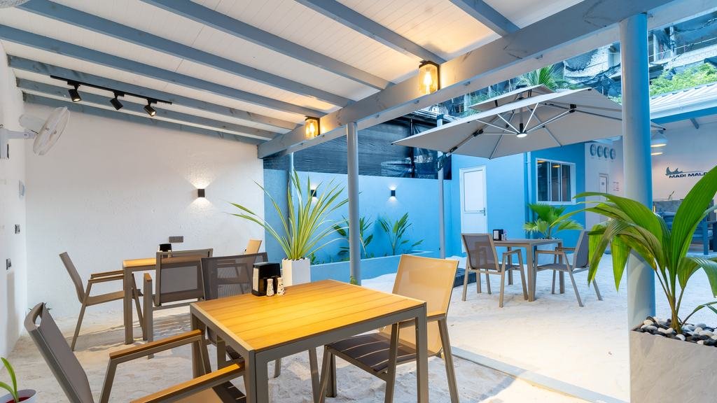 The courtyard at Madi: tables and chairs under a pitched roof and a parasol, planted pots along the blue and white walls.